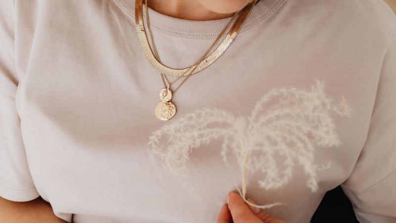 Woman in a beige tee holding pampas grass, layered gold chains and coin pendants.