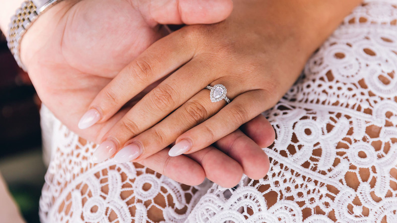 Couple's hands on white lace, she wears a pear-shaped diamond halo engagement ring.