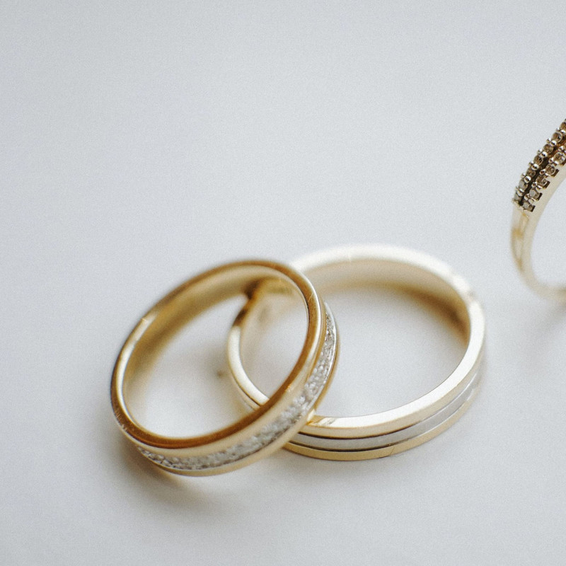 Two gold wedding bands, one channel-set with small stones, on a plain pale surface (engagement ring mostly cropped out at the edge).