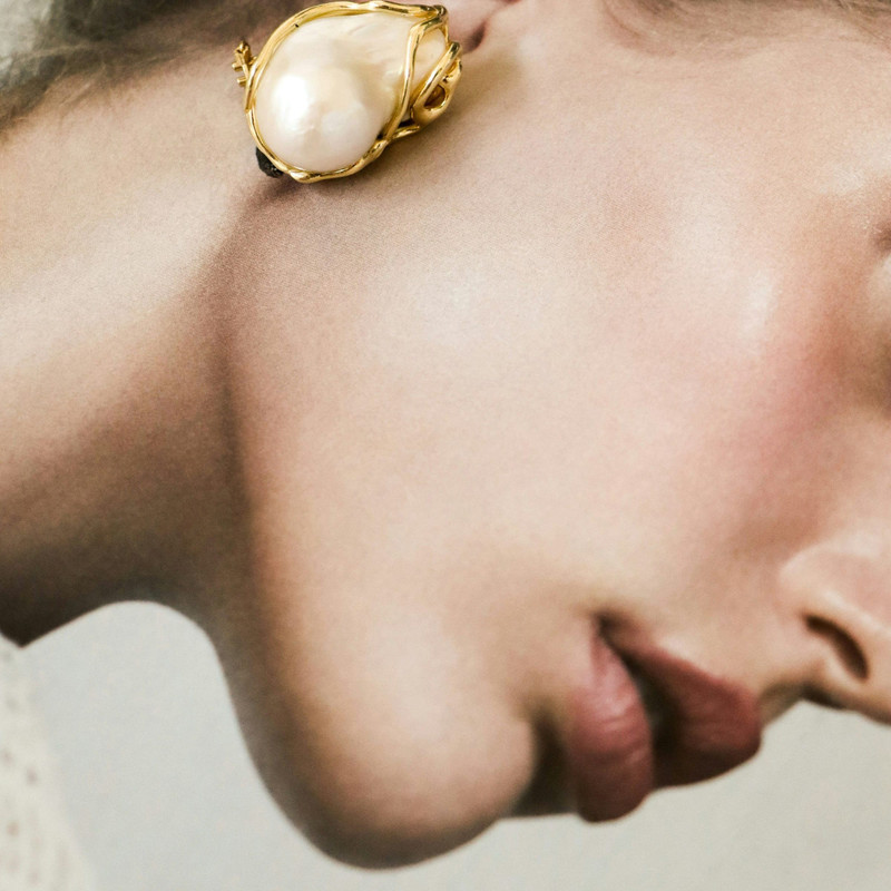 Close crop of an ear wearing a baroque pearl earring wrapped in gold, soft warm skin.