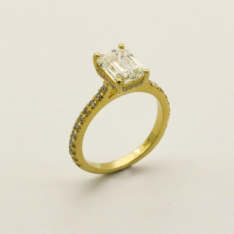Yellow-gold emerald-cut diamond solitaire with pavé shoulders, angled on a plain white background.