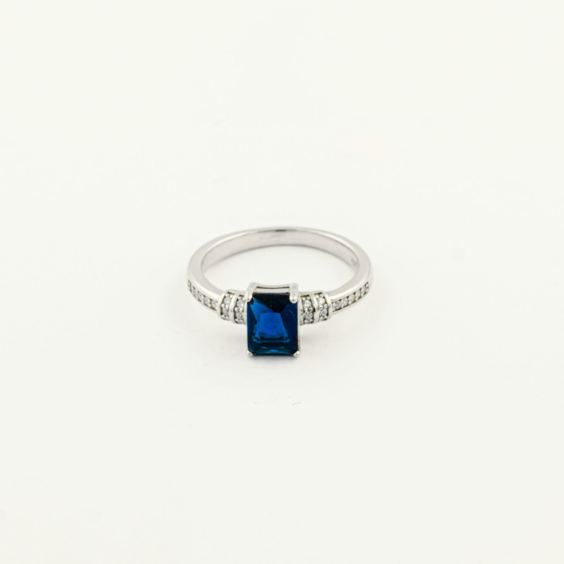 White-gold ring with an emerald-cut blue sapphire flanked by diamond shoulders, on white.