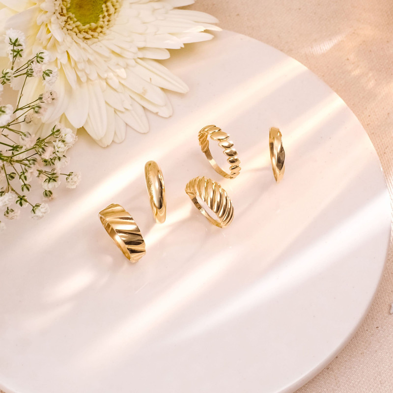 Set of twisted and ribbed gold rings on a white plate with soft window light and a white daisy.