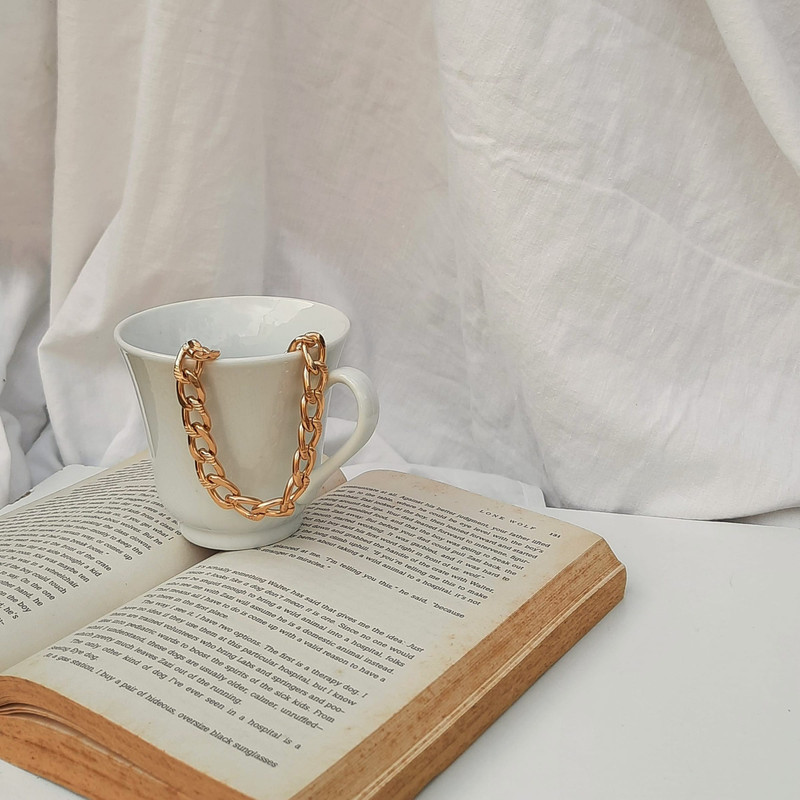 Chunky gold chain looped over a white mug on an open book, white drapery behind.