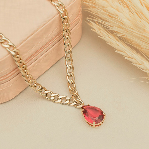 Gold curb chain with a red teardrop stone on a beige box with pampas grass.