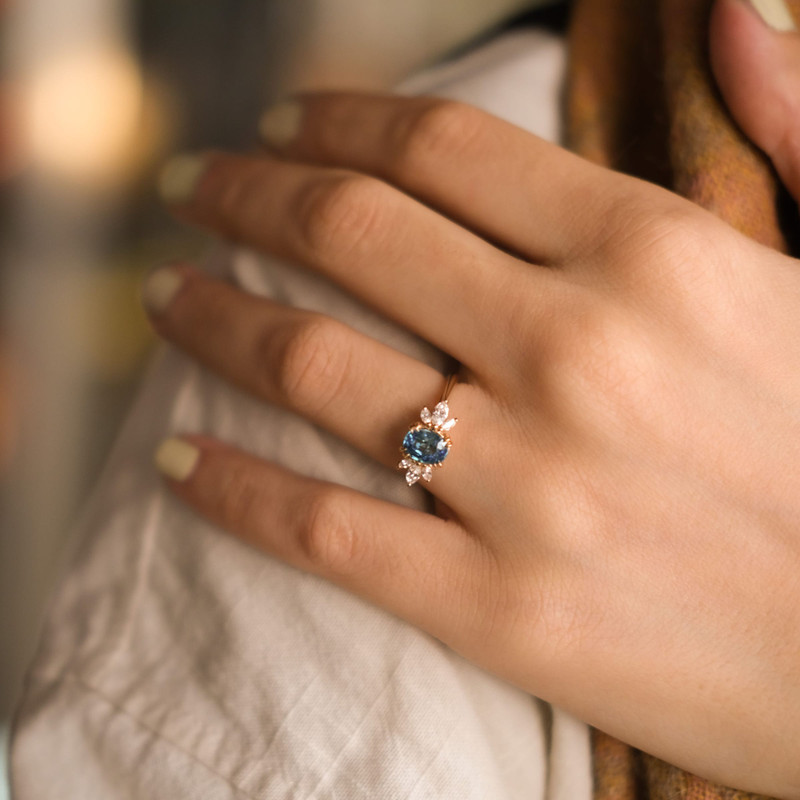 Hand resting on linen wearing a rose-gold ring with a blue stone and diamond accents.