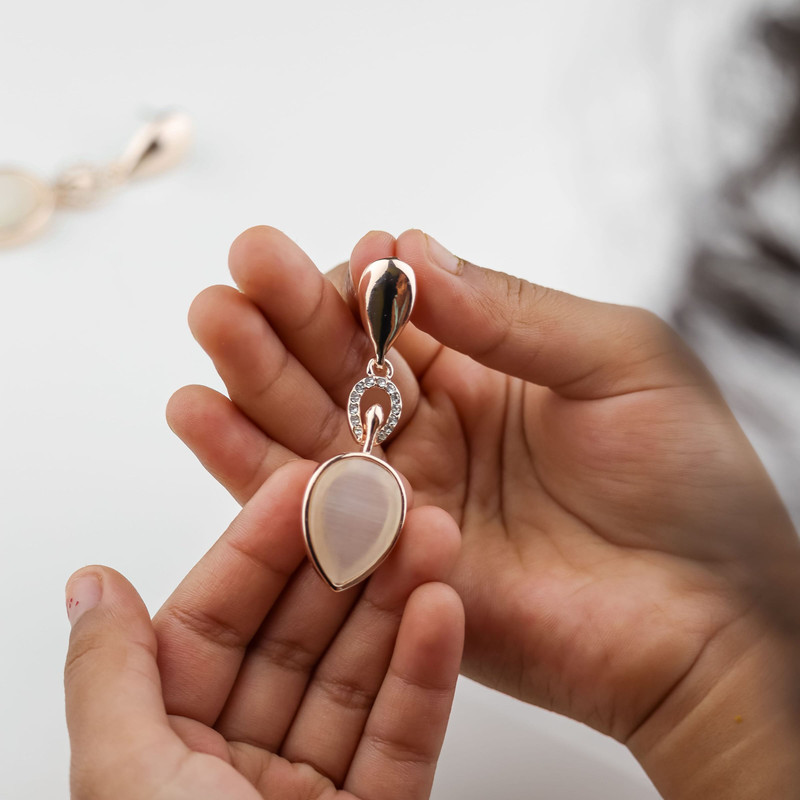 Hands holding a rose-gold teardrop pendant with a pale pink stone, soft white light.