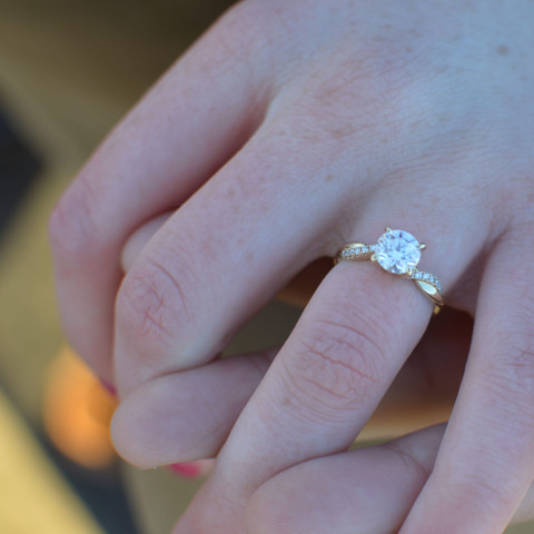 Macro of a round solitaire with pavé band on a finger, soft pink light.
