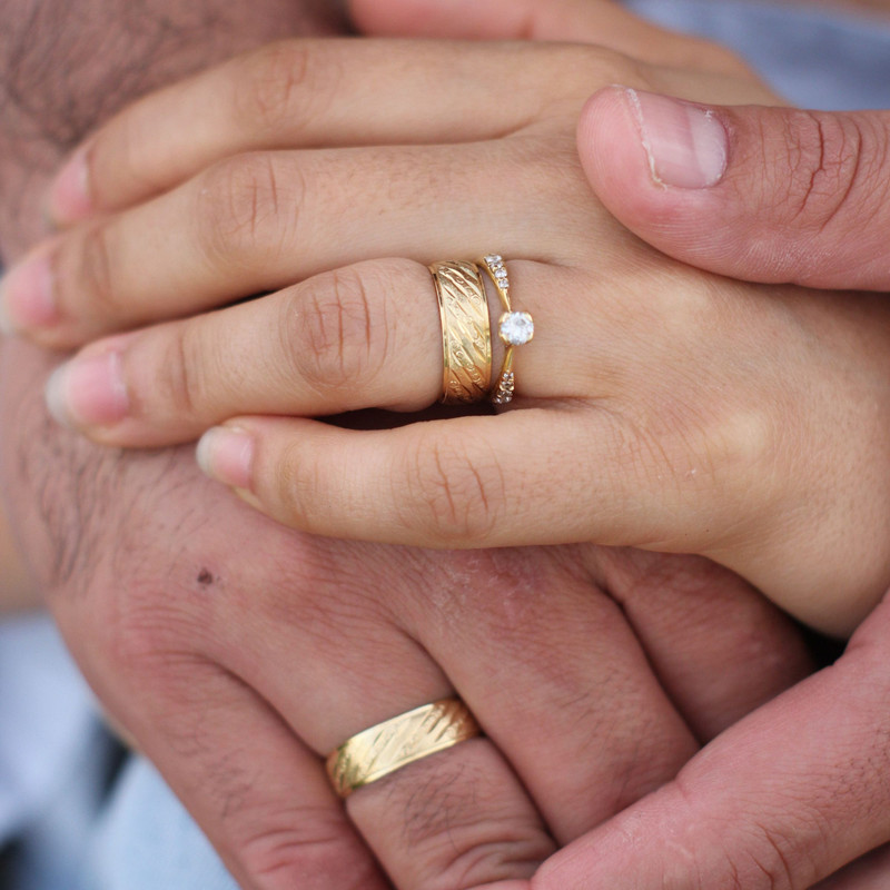 Couple's hands, she wears a gold wedding band with a diamond engagement ring, warm skin tones.