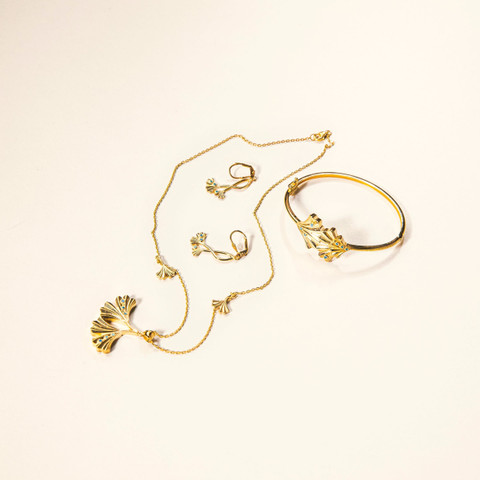 Gold necklace with ginkgo-leaf charms laid out with matching bangle and earrings on white.