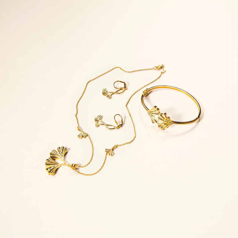 Gold necklace with ginkgo-leaf charms laid out with matching bangle and earrings on white.
