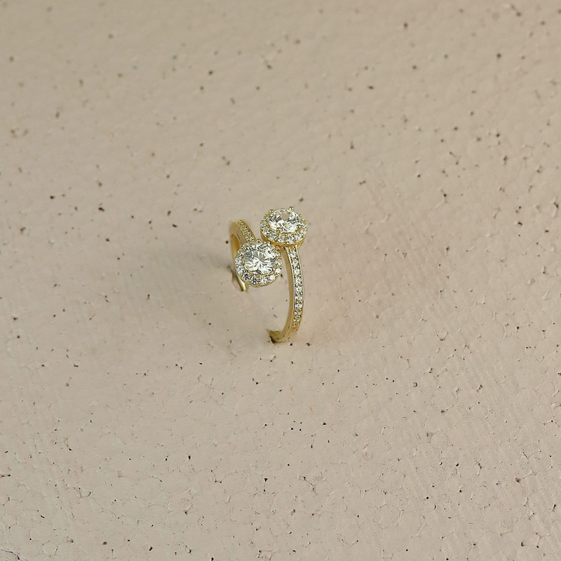 Same two-stone halo ring standing on a warm sand-coloured speckled surface.
