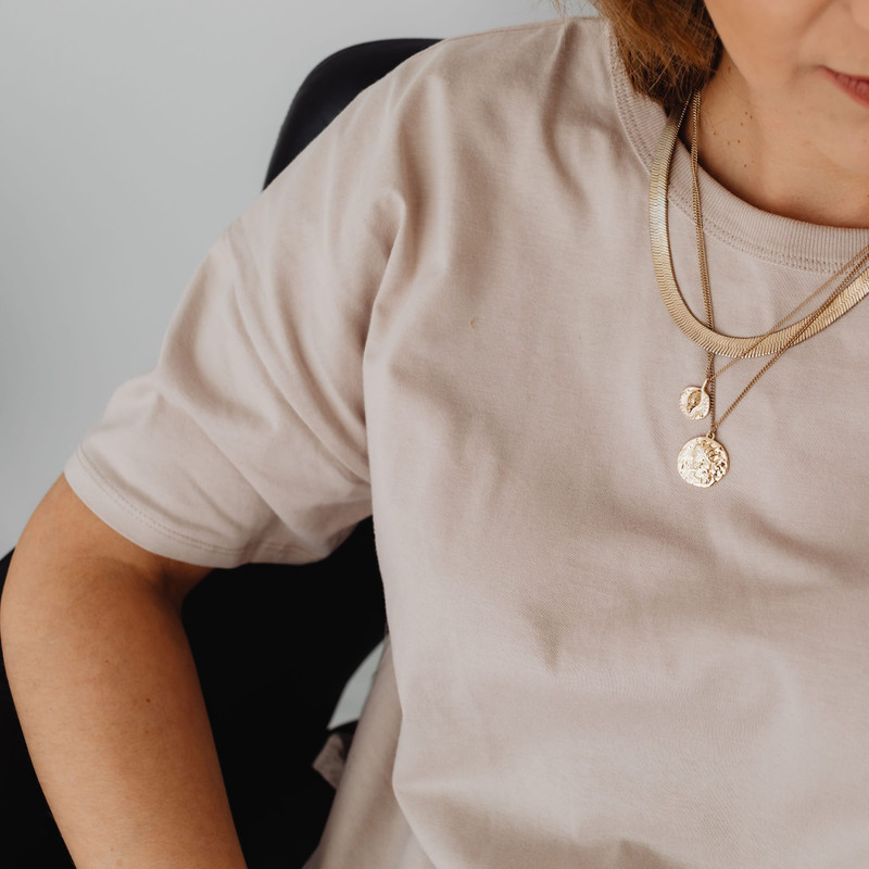 Woman in a beige tee wearing layered gold chains with coin pendants.