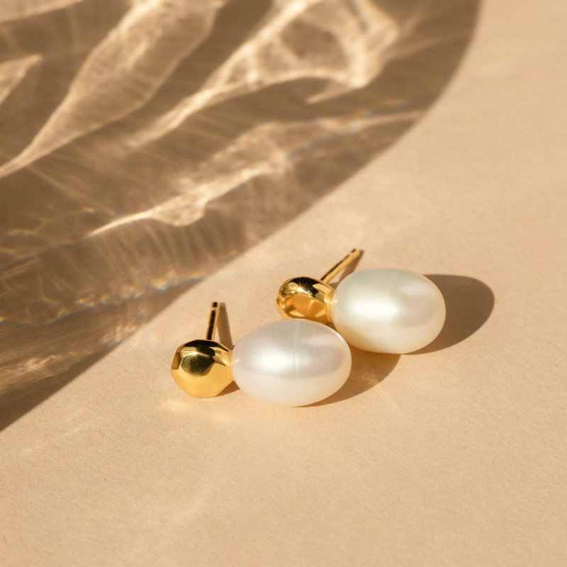 Freshwater pearl drop studs and a gold ball stud on warm sand with rippling light shadows.