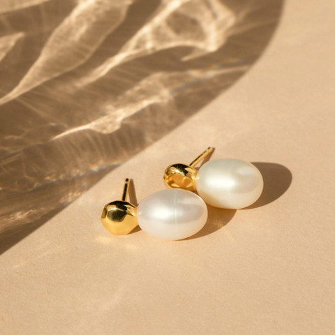 Freshwater pearl drop studs and a gold ball stud on warm sand with rippling light shadows.