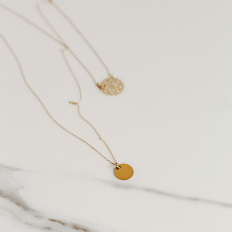 Two fine gold necklaces with filigree and disc pendants laid on white marble.