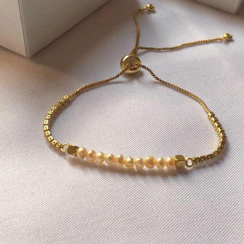 Gold slider bracelet with a row of small pearl beads, in a white box on blush fabric.