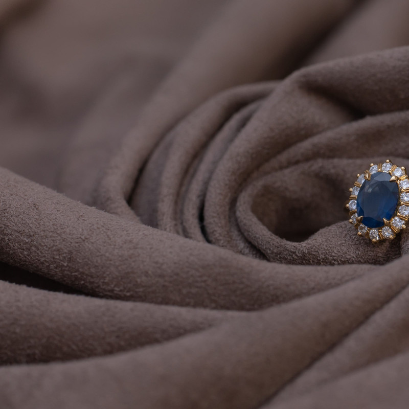 Gold ring with an oval sapphire in a diamond halo nestled in taupe suede.