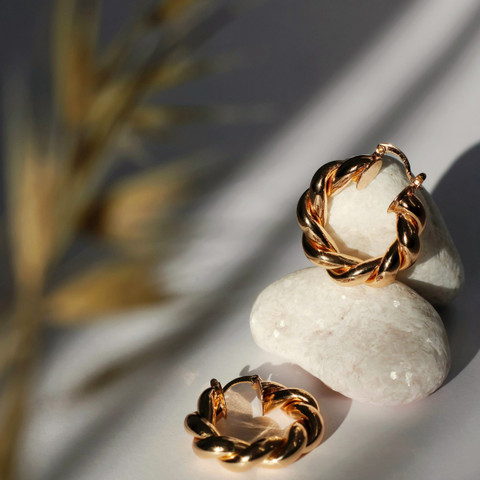 Two twisted rope-style gold rings on smooth white pebbles, soft shadow and dried grass blur.