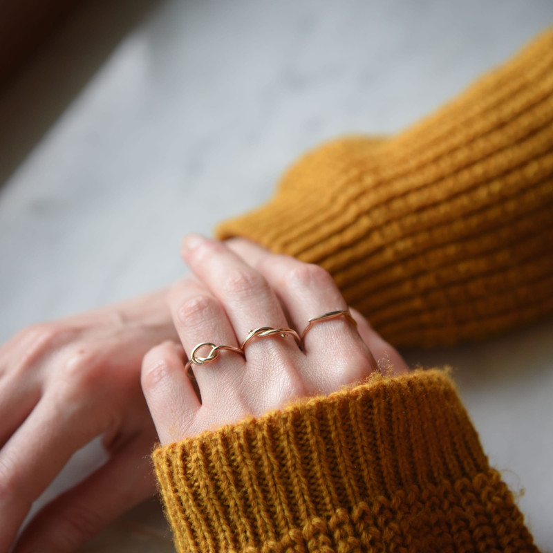 Hands in a mustard knit sleeve wearing slim stacked gold rings on a pale marble surface.